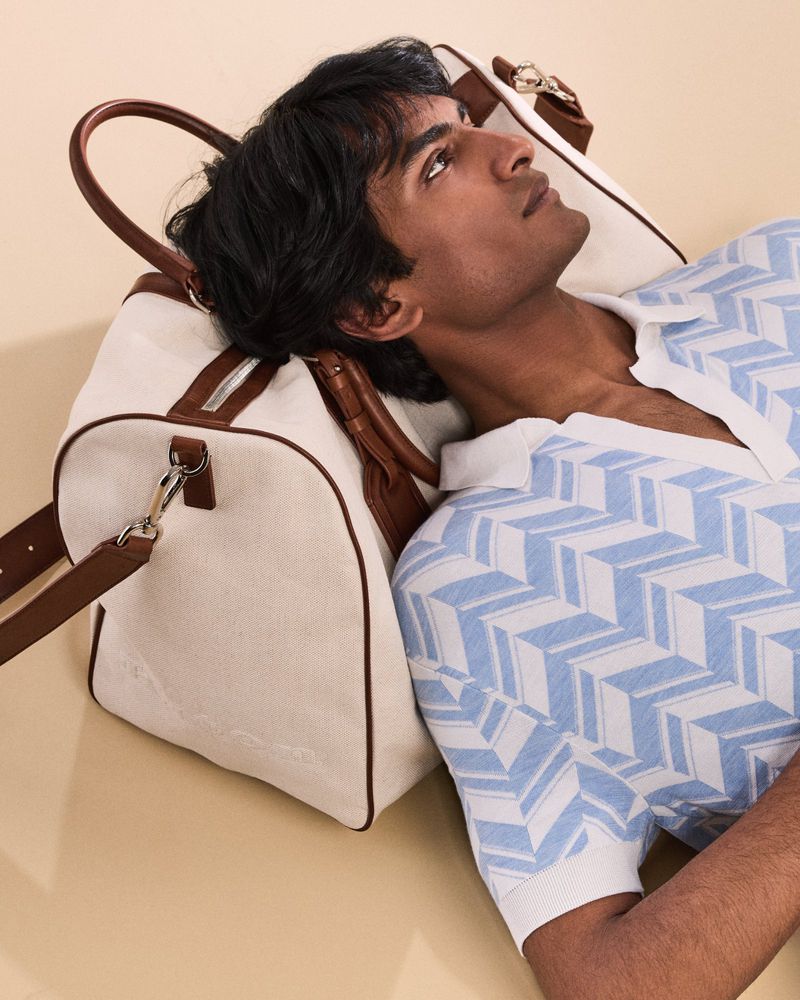An adult male rests near a cream-colored duffel bag with brown leather accents, wearing a blue and white patterned collared shirt.