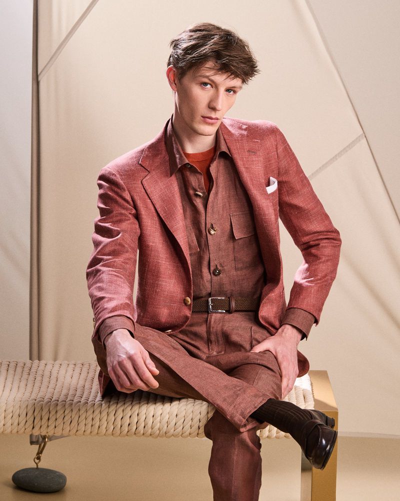 A stylish young man is sitting, wearing a rust-colored suit with a brown shirt and black shoes, posing in a studio setting. 