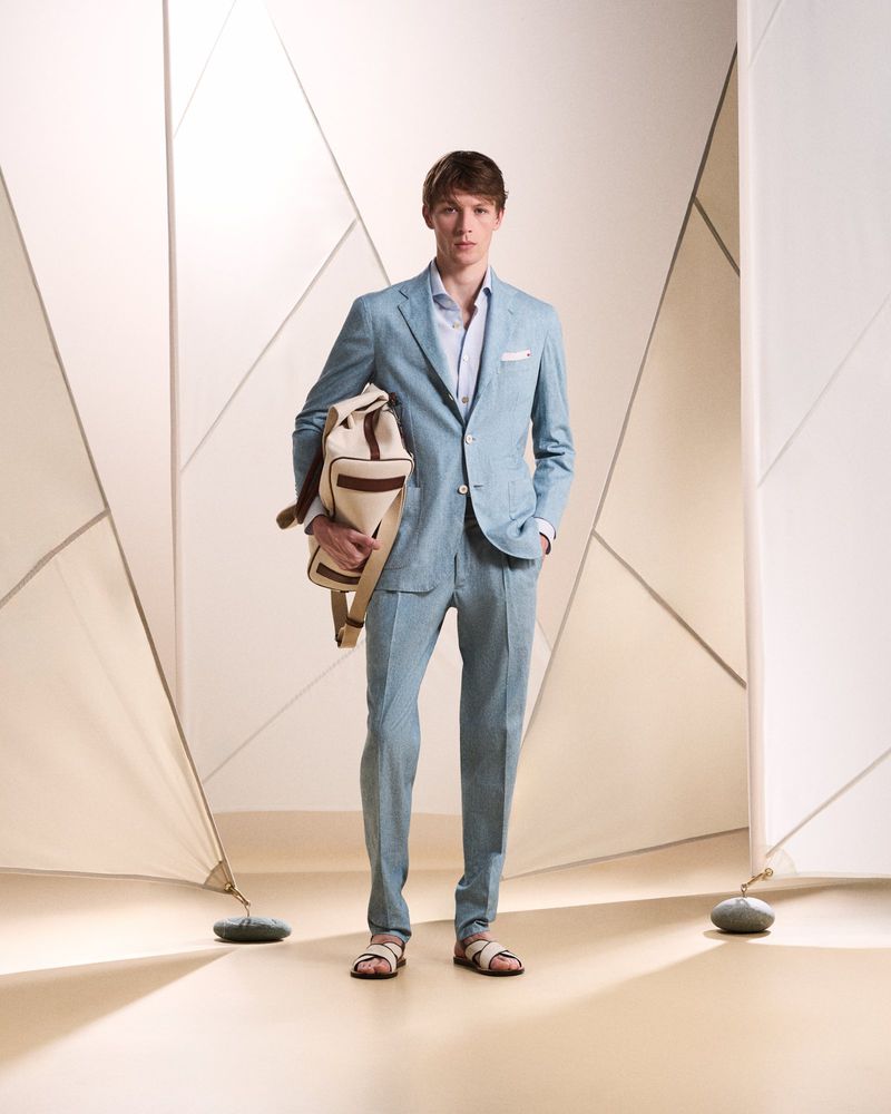 A young adult man in a light blue suit stands in a studio, carrying a duffel bag. He's wearing sandals and is well-lit against a geometric backdrop.
