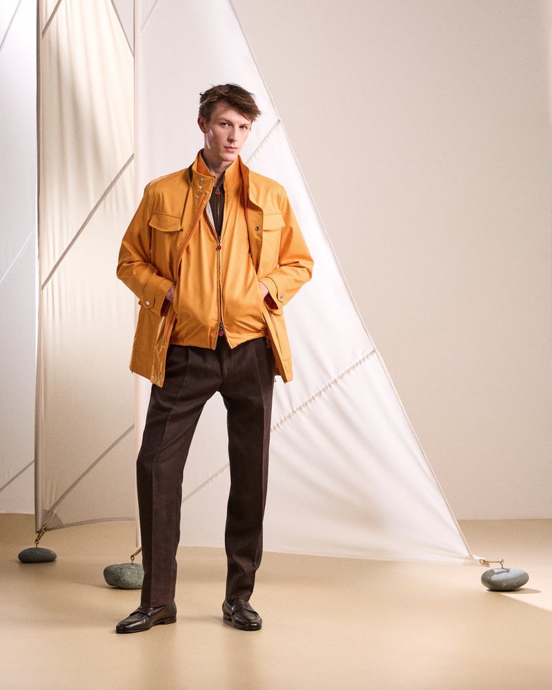 Fashionable adult male model poses in an ochre jacket and brown trousers in a studio with a sail prop.