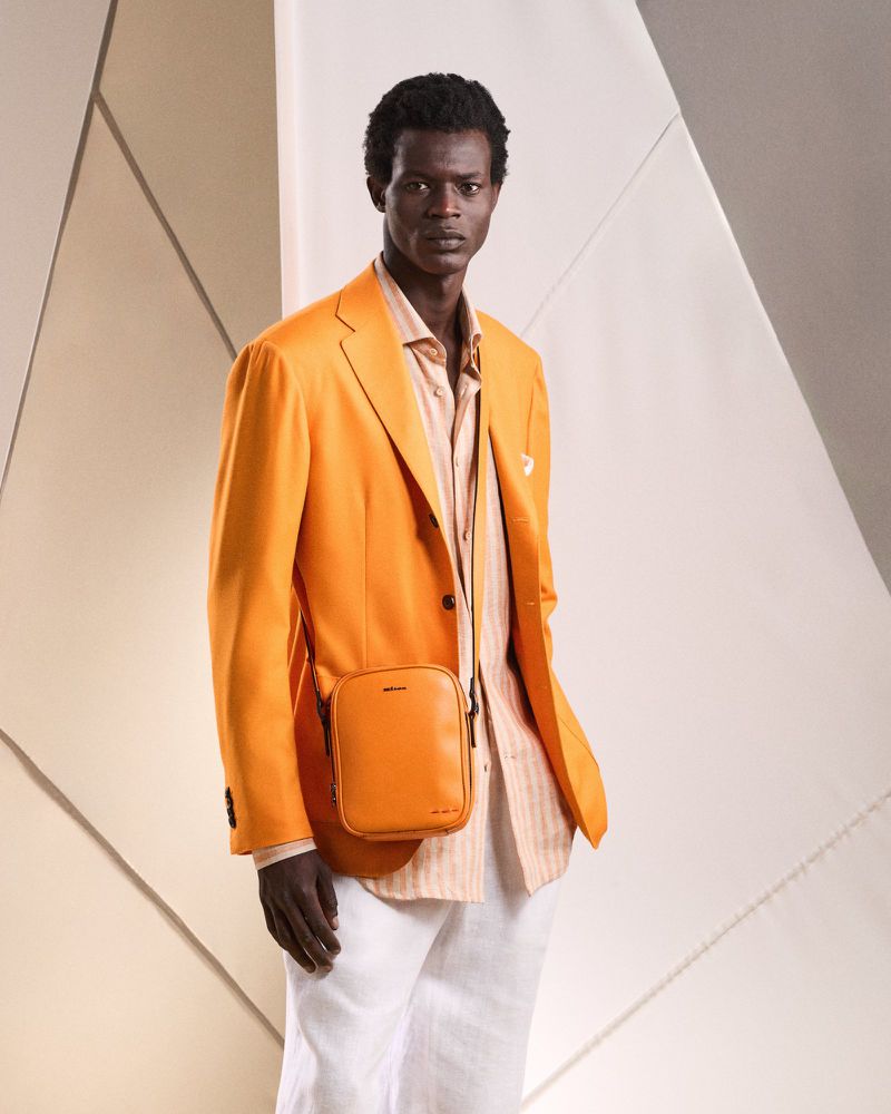 Portrait of dark-skinned adult male in an orange blazer and white pants, wearing a matching orange crossbody bag against geometric background.