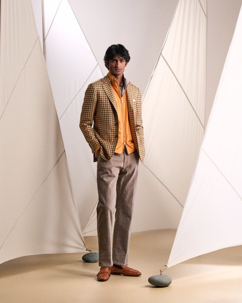 Full-length shot of a dark-haired man in an orange shirt and brown suit, posing in front of a geometric white backdrop.