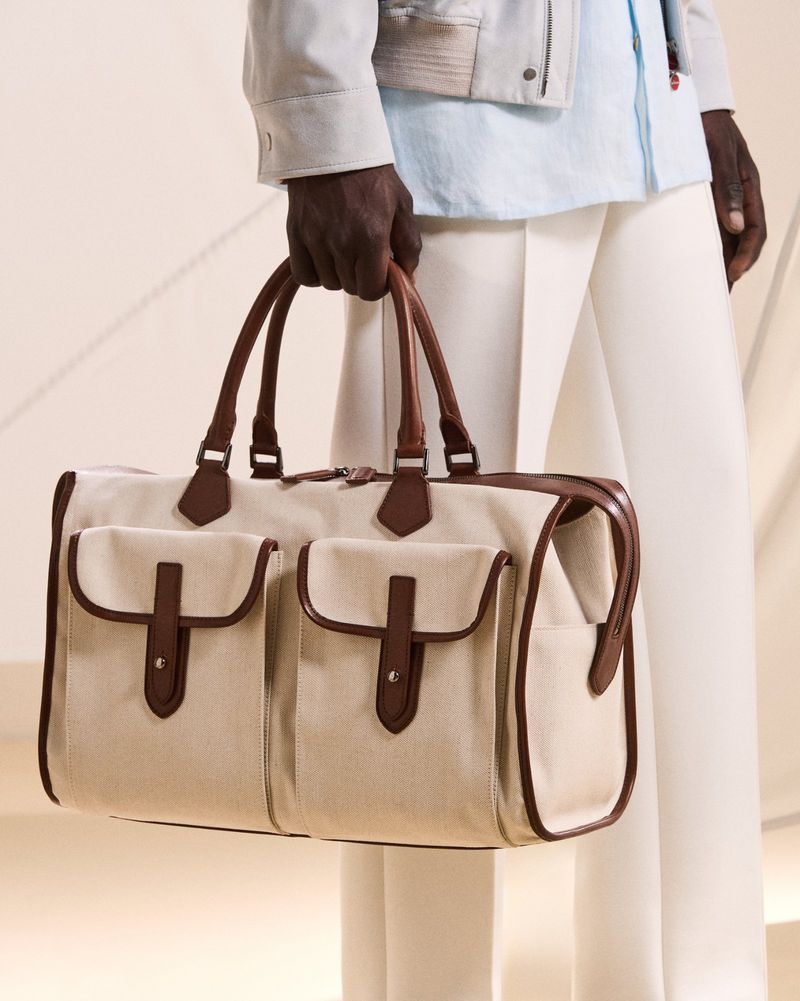 Canvas and leather duffel bag held by an adult. The person is wearing white pants and light blue shirt. The bag is tan and brown.