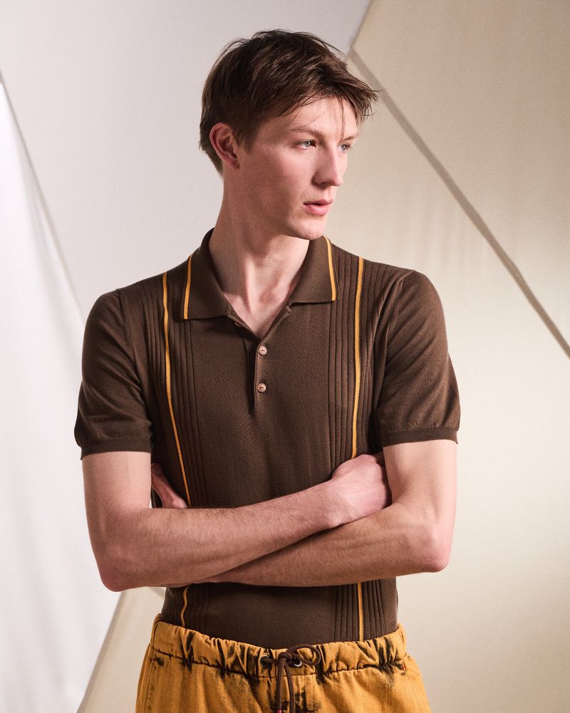 Portrait of a light-skinned young adult male in a brown, short-sleeved polo shirt with yellow trim, posing indoors with arms crossed.
