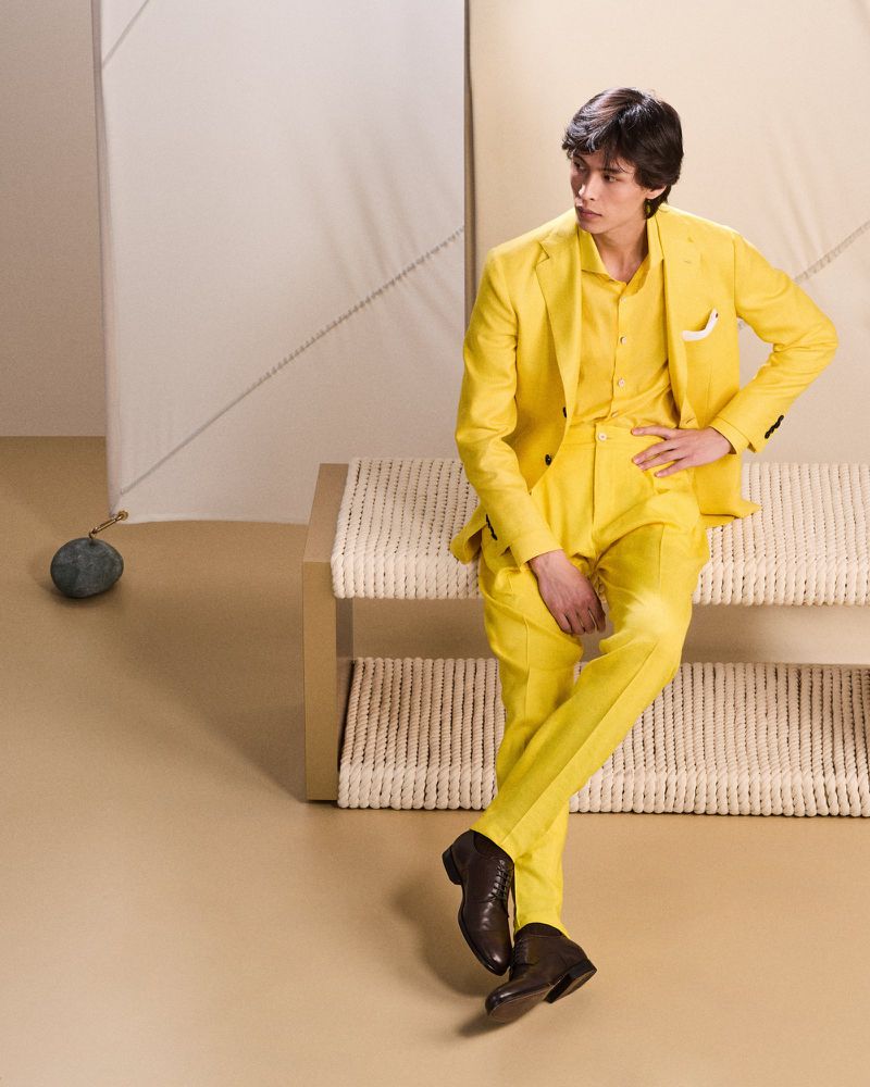 A young adult male poses seated on a woven bench wearing a yellow suit, yellow shirt, and brown leather shoes in a studio setting.