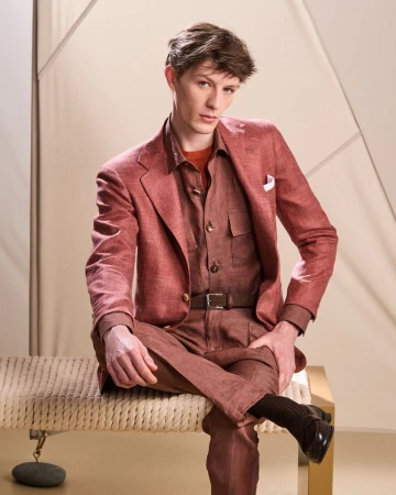 Model Finnlay Davis seated in a dusty rose tailored linen suit and brown shirt on a woven bench for Kiton.