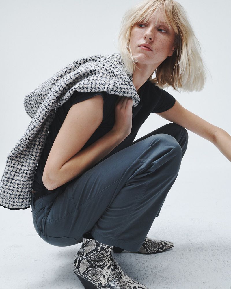 Blonde woman poses crouching down in a black top, grey-green pants, patterned boots, and houndstooth jacket in a studio setting.