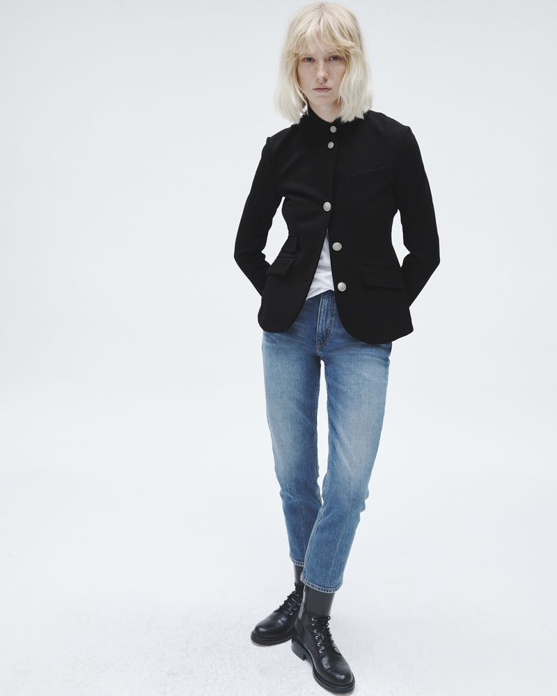 A woman with blonde hair stands in front of a white backdrop wearing a black jacket, blue jeans, and black boots in a studio shot.