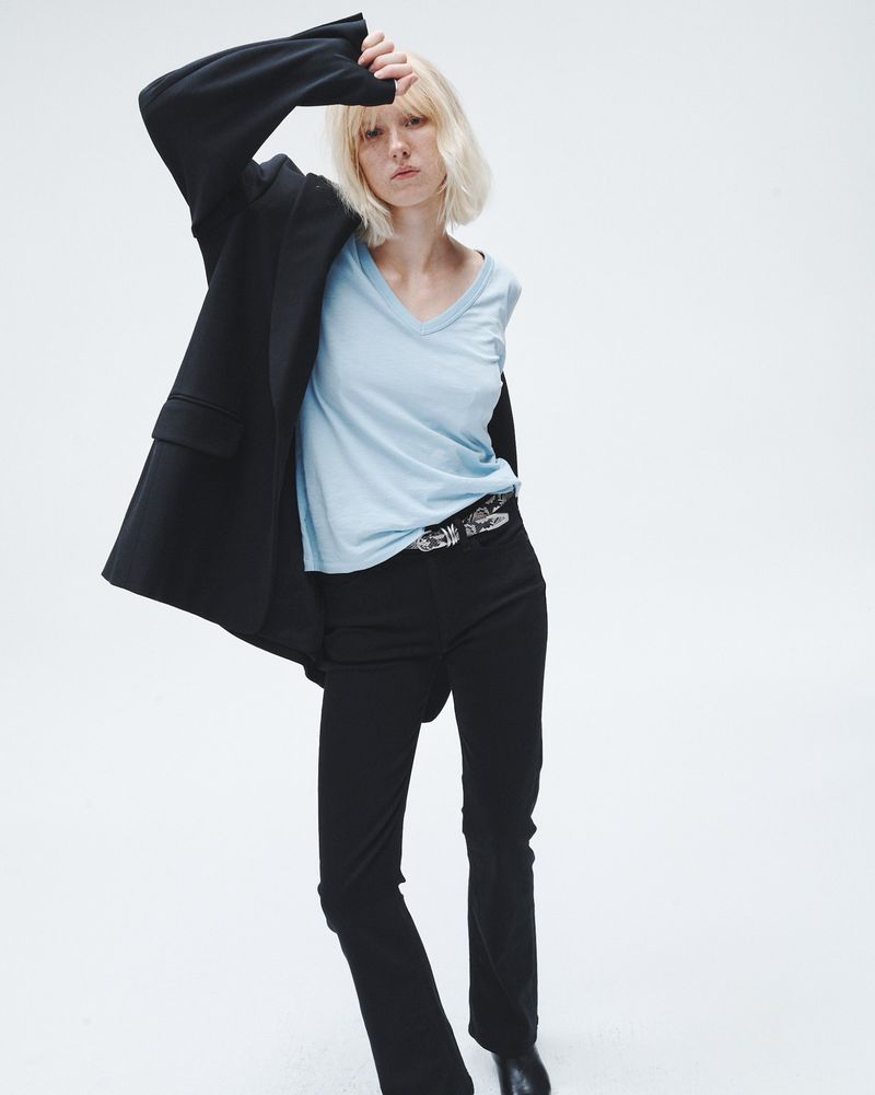 A young woman with blonde hair poses in a studio with a black blazer, light blue t-shirt, black pants, and black shoes, with a white background.
