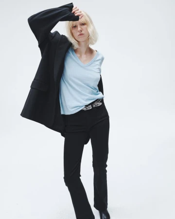 A young woman with blonde hair poses in a studio with a black blazer, light blue t-shirt, black pants, and black shoes, with a white background.