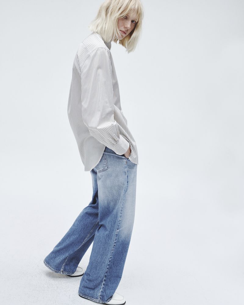 A blonde-haired person in a striped shirt and faded jeans poses against a white backdrop in a fashion shot with a shallow depth of field.