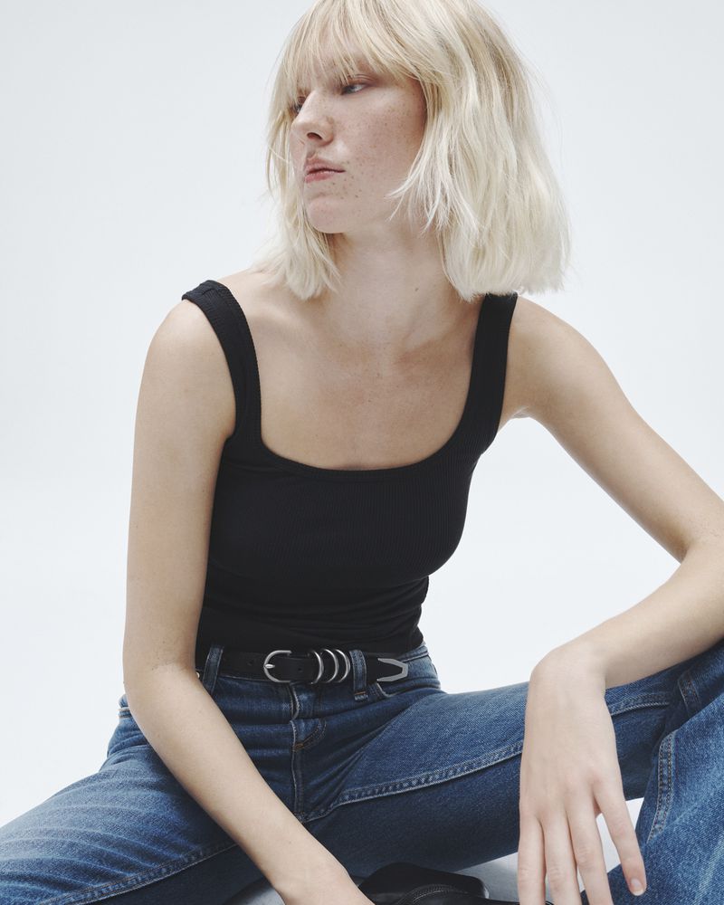 A woman with blonde hair poses in a black tank top and blue jeans against a white background in a studio portrait.
