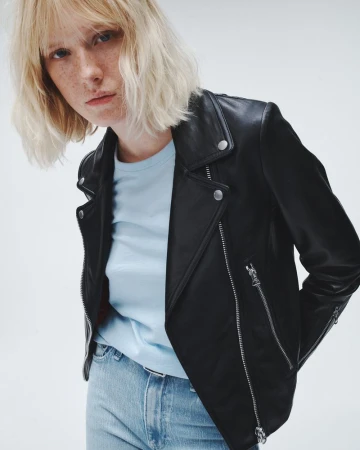 A blonde woman poses in a black leather jacket, light blue tee, and jeans against a white backdrop, showcasing fashion and style.