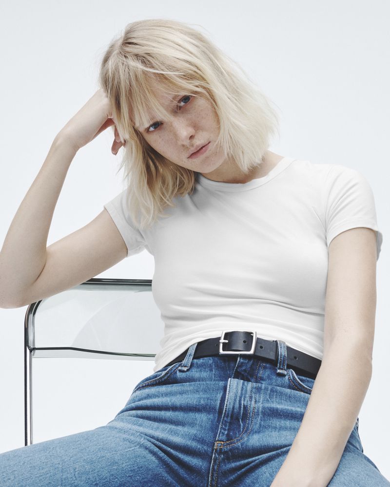 Young woman with blonde hair poses in a casual white T-shirt and blue jeans, seated in a clear chair against a bright white backdrop.