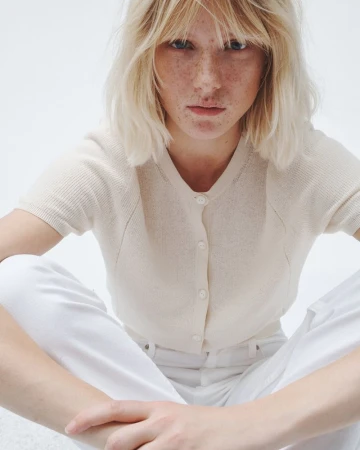 A young woman with blonde hair poses in a bright studio wearing a cream-colored cardigan and white pants, shot with a shallow depth of field.