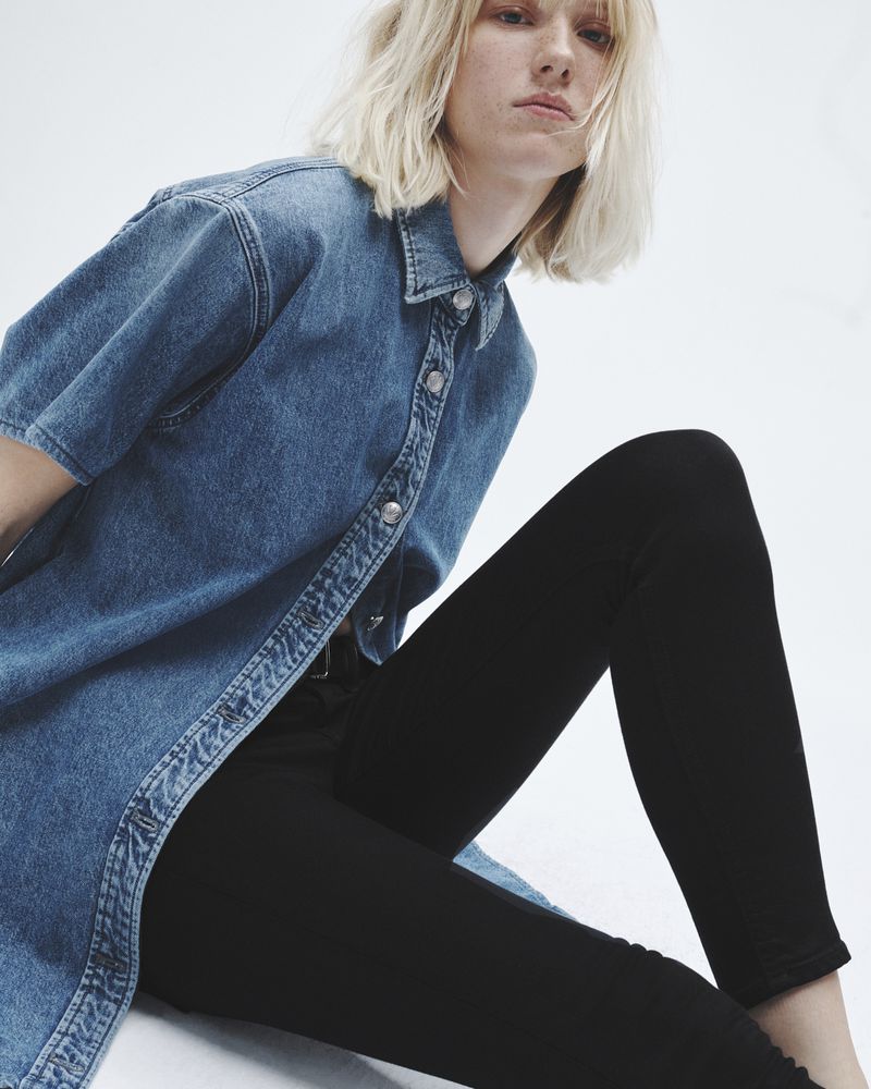 A young adult woman with blonde hair poses in a blue denim shirt-dress and black leggings on a white background.