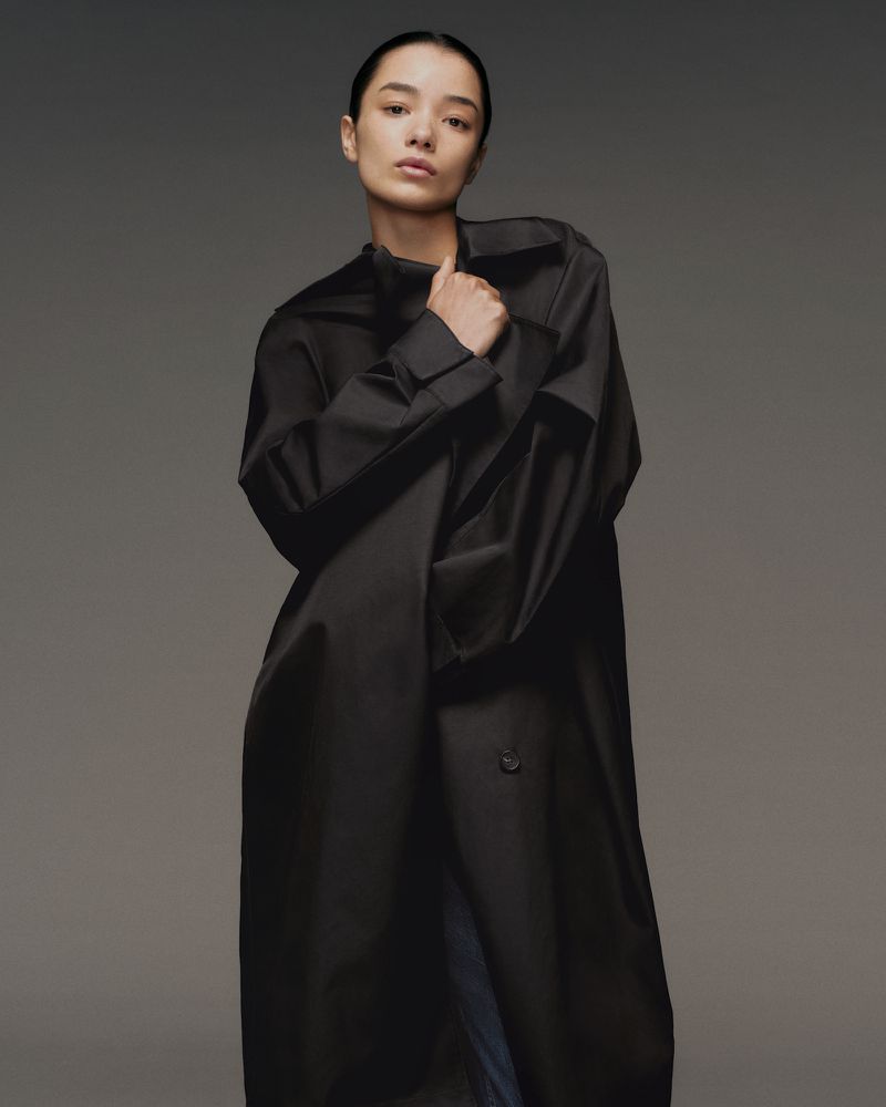Model wearing an oversized black trench coat with slicked-back hair clutching the lapel against a neutral grey studio backdrop.