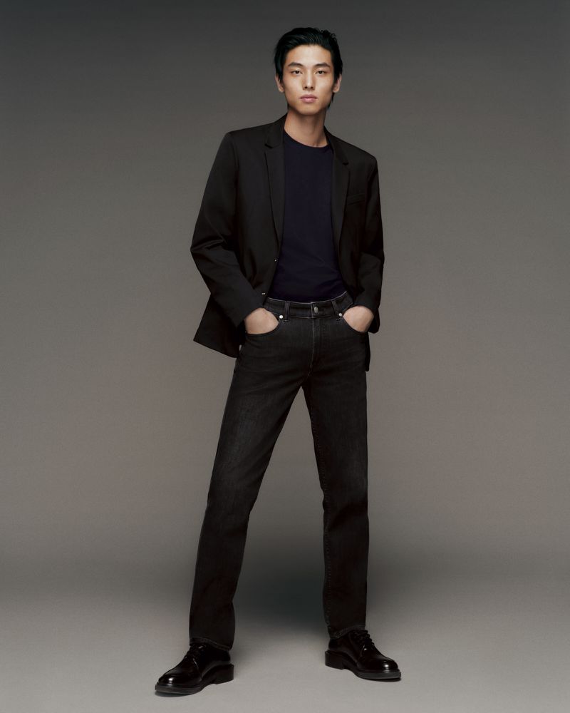 Full-length portrait of an Asian adult male wearing a blazer, dark top, and dark jeans, standing in a photo studio with a grey backdrop.