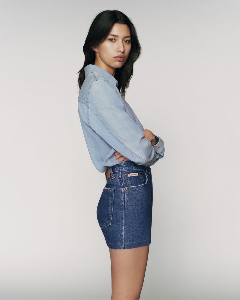 Standing woman with arms crossed in denim shirt and skirt against a white backdrop in a fashion forward full shot.