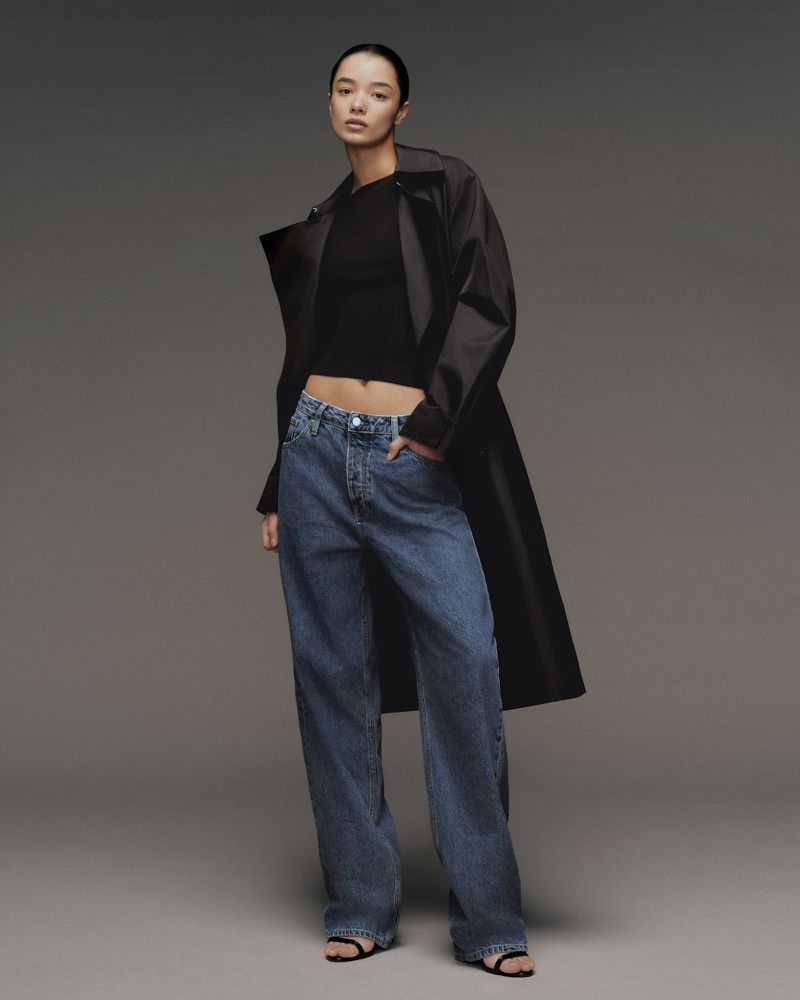 A woman poses in blue jeans, a black top, and a dark coat against a gray background, showcasing a modern and casual fashion style.