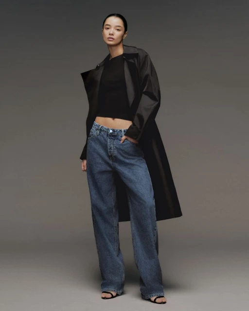 A woman poses in blue jeans, a black top, and a dark coat against a gray background, showcasing a modern and casual fashion style.
