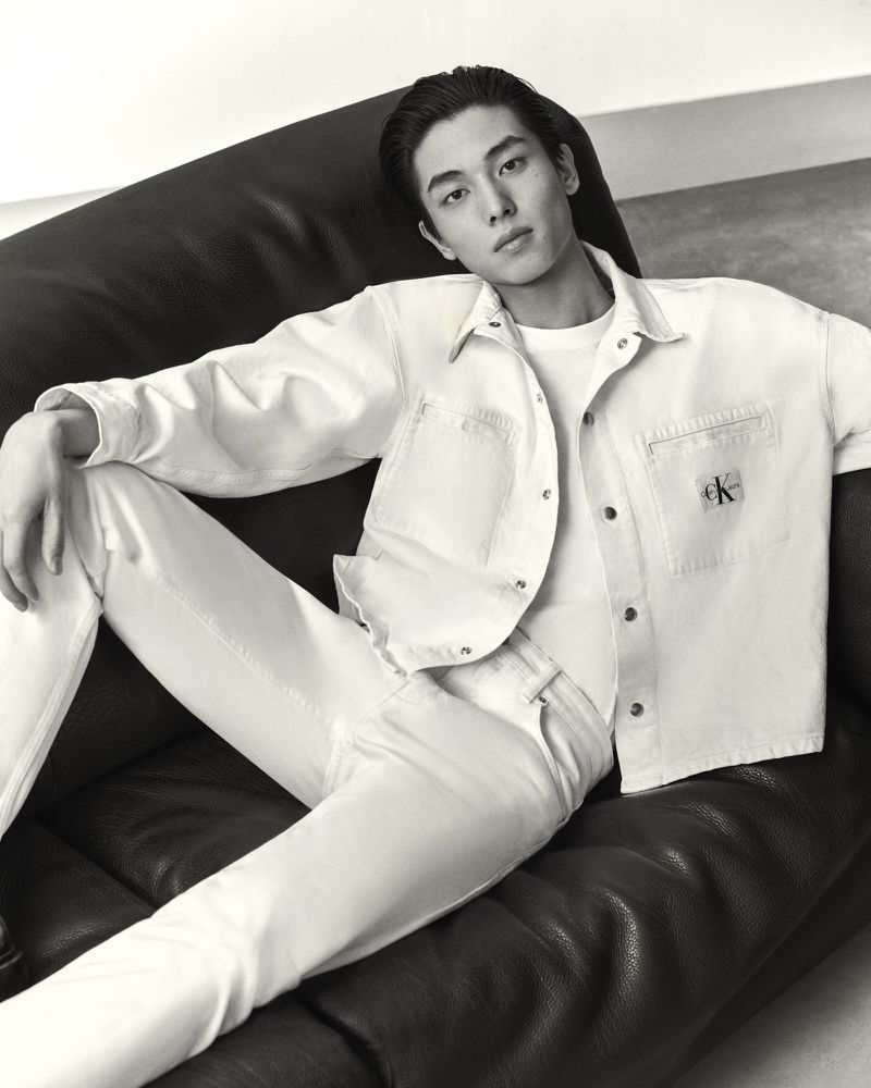 Black-and-white portrait of a young Asian man reclining on a dark leather couch wearing a white denim jacket and white jeans.