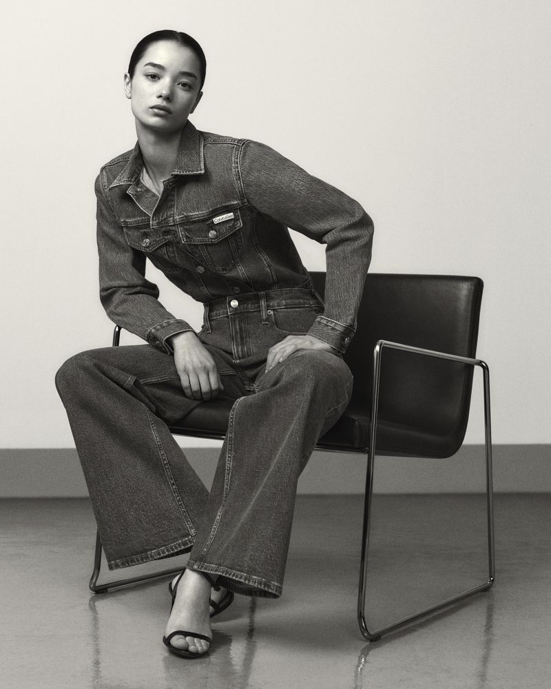 A seated model wearing a matching denim jacket and wide-leg jeans in an armless black chair against a neutral studio backdrop.