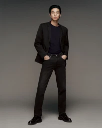 Full-length portrait of a male model in a black blazer, dark t-shirt, and dark jeans against a neutral grey studio backdrop.