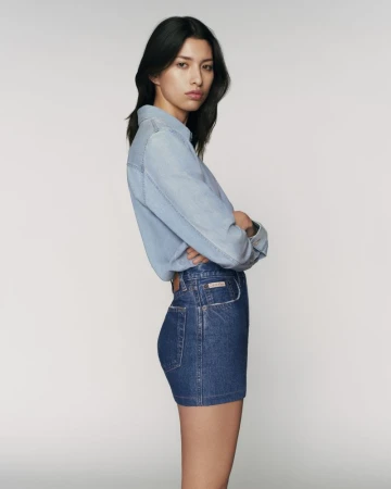 East Asian model in a light blue denim shirt and dark denim mini skirt standing in profile against a neutral grey studio backdrop.
