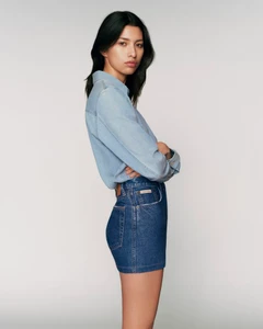 East Asian model in a light blue denim shirt and dark denim mini skirt standing in profile against a neutral grey studio backdrop.