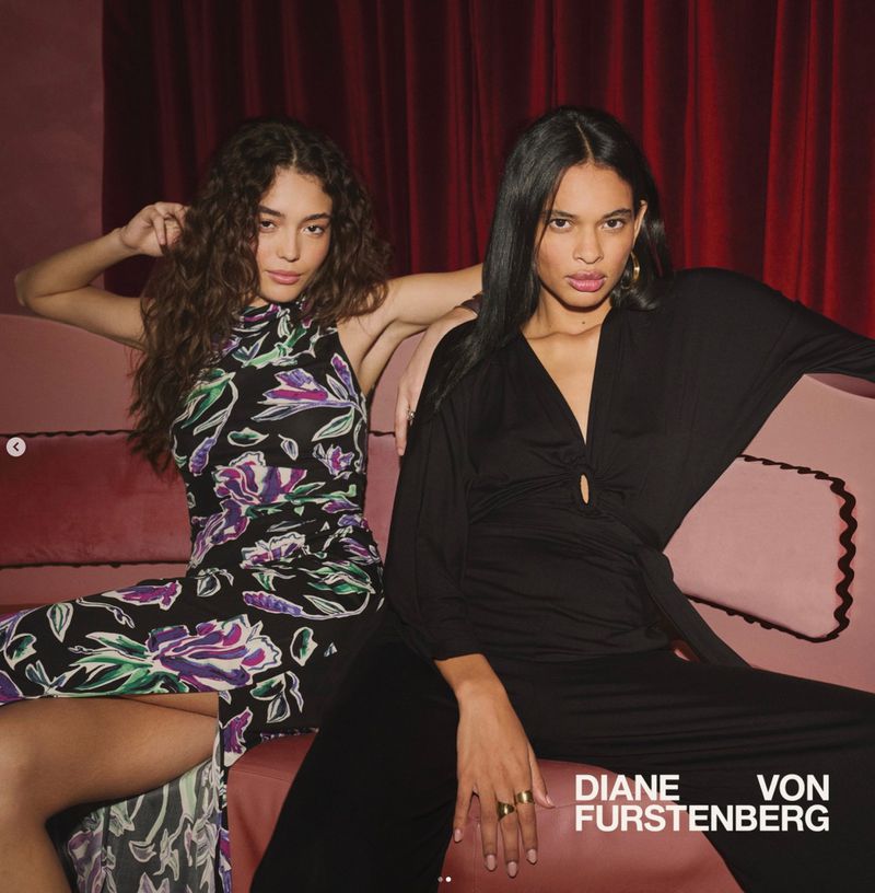 Two fashion models wear black and floral print jumpsuits while posing on a pink couch in front of a maroon curtain backdrop.