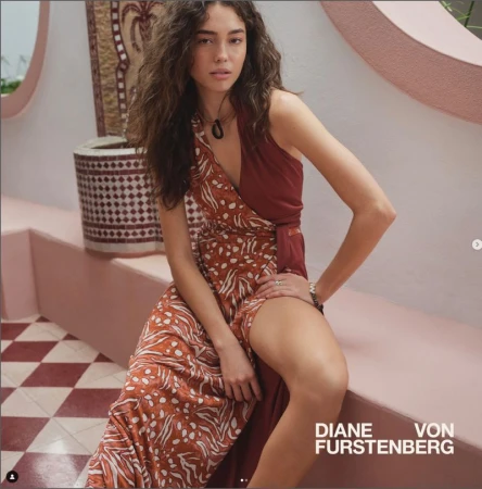 Woman wearing patterned wrap dress posing in an indoor setting with patterned tile floor and light-colored architectural details in the background.