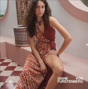 Model in a printed terracotta wrap dress seated on a pink bench beside a tiled fountain in an outdoor courtyard.