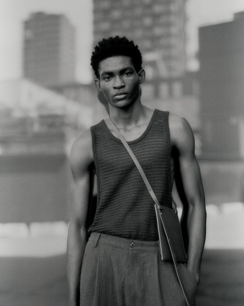 Monochrome portrait of a Black man wearing a textured knit tank top, pleated trousers, and a slim crossbody bag outdoors.