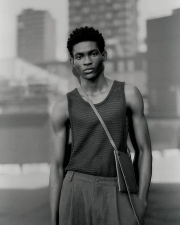 Monochrome portrait of a Black man wearing a textured knit tank top, pleated trousers, and a slim crossbody bag outdoors.