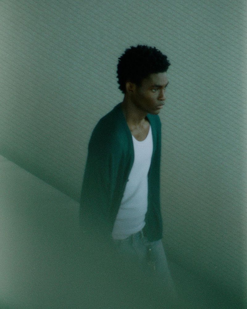A young adult Black male is shown waist-up, wearing a white t-shirt, green cardigan, and light jeans. He's indoors with a textured wall behind.