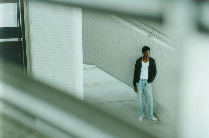 An adult male model with dark hair stands in a neutral-toned indoor space wearing jeans, a white t-shirt, and a dark cardigan.