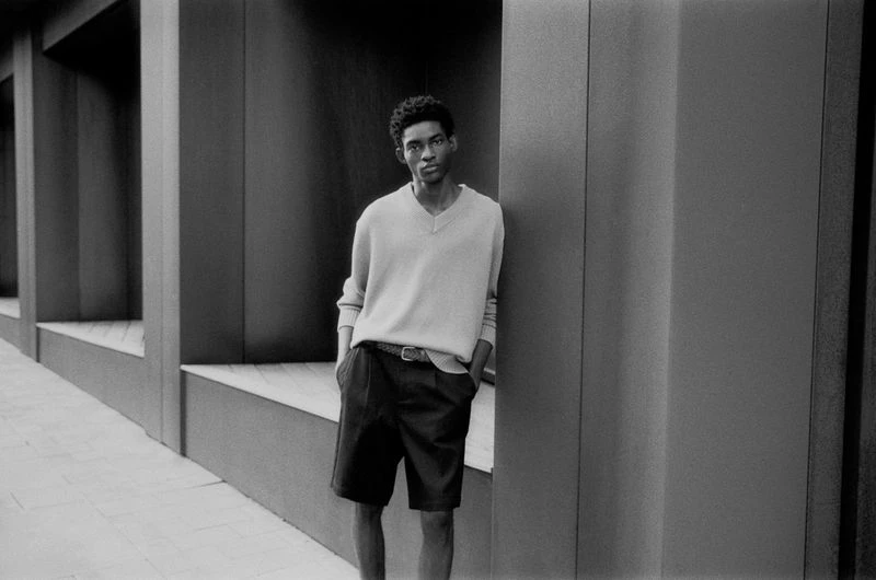 A slim, dark-skinned male model in a v-neck sweater and dark shorts poses against a modern architectural backdrop, bathed in soft, diffused daylight.