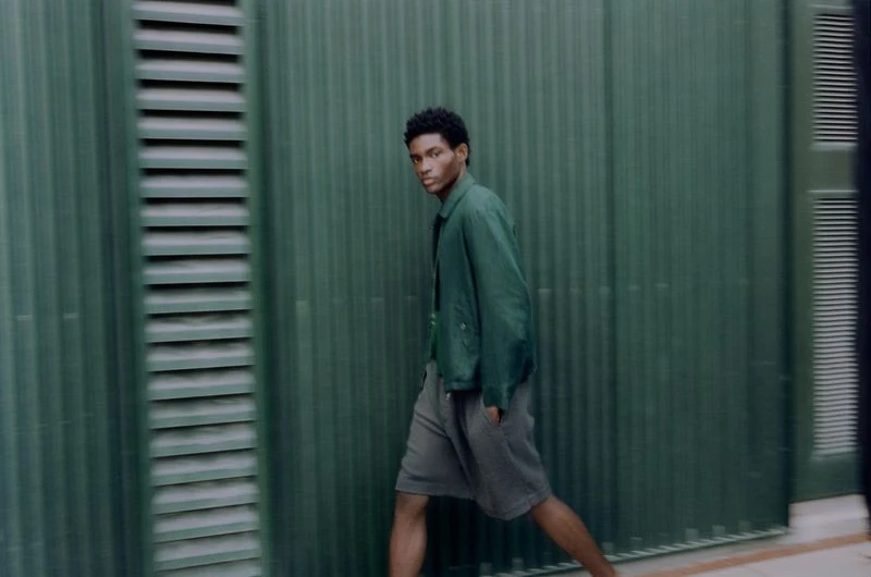 A young adult man walks in front of a green corrugated wall, wearing a matching green jacket and gray shorts in an urban setting.