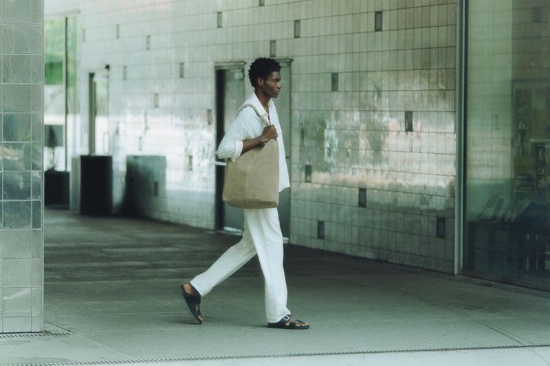 A person walks in an urban outdoor setting with a tiled wall, wearing casual white clothing and carrying a brown tote bag on their shoulder.