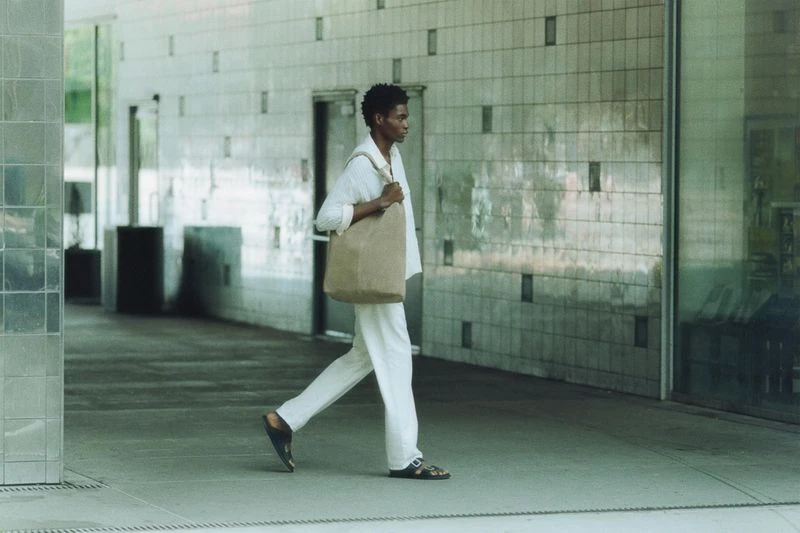 A person walks in an urban outdoor setting with a tiled wall, wearing casual white clothing and carrying a brown tote bag on their shoulder.