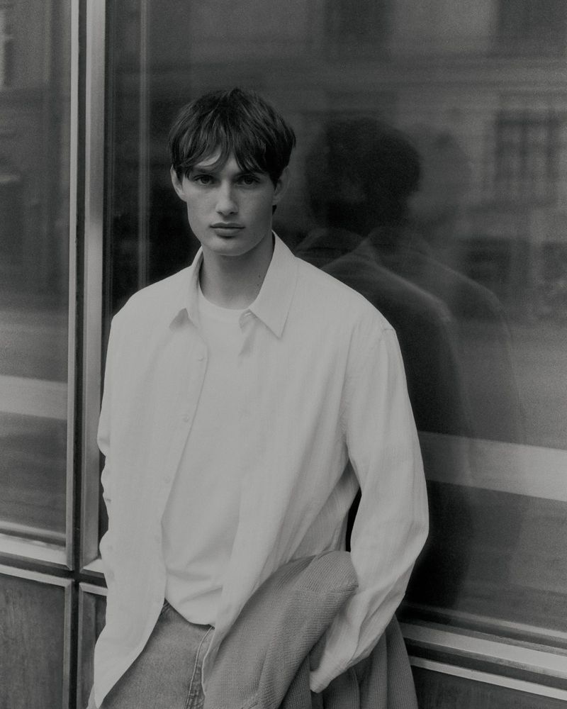 Monochrome medium shot of a male model with brown hair, wearing a white button-down shirt and jeans, posing against a glass building.