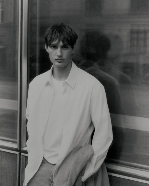 Monochrome medium shot of a male model with brown hair, wearing a white button-down shirt and jeans, posing against a glass building.