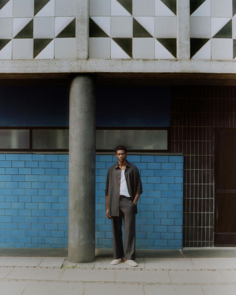 Man in stylish gray shirt and pants posing against a blue tiled wall next to a concrete column in an outdoor, urban setting.