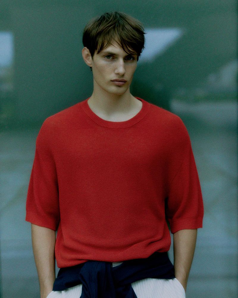 Portrait of a young adult man wearing a red short-sleeved knit shirt, gazing at the camera with a neutral expression, against a blurry backdrop.