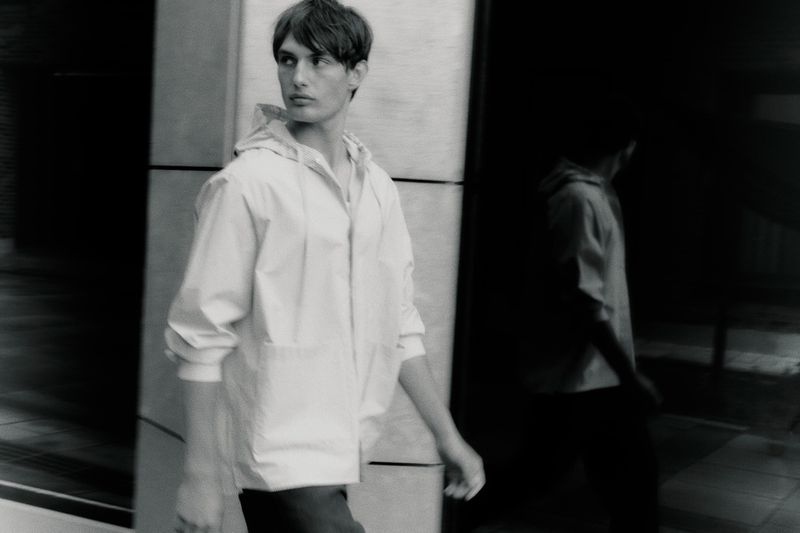 Monochrome shot of a man wearing a light hooded jacket walking past a modern building exterior with a glass reflection.