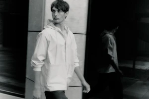 Monochrome shot of a man wearing a light hooded jacket walking past a modern building exterior with a glass reflection.