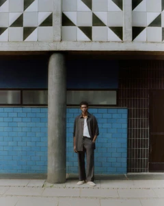 Evans Ikechukwu in a relaxed dark grey matching set standing by a concrete pillar and blue tile wall for Zara Man.