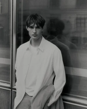 Young male model wearing an unbuttoned white shirt and t-shirt standing against a glass storefront reflection.