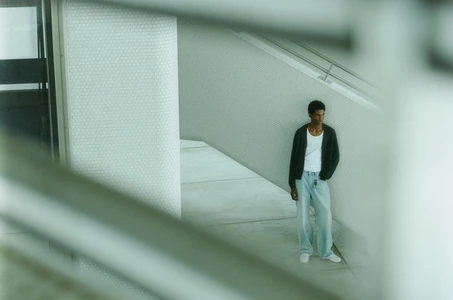 A man wearing a dark jacket over a white tank top and light jeans standing near modern architectural walls.